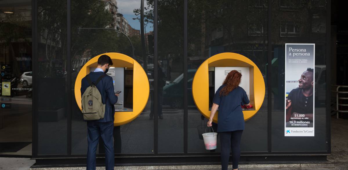 Una señora limpia un cajero de CaixaBank mientras un chico realiza una operación en la calle Orense (Madrid)