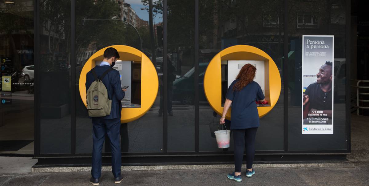 Una señora limpia un cajero de CaixaBank mientras un chico realiza una operación en la calle Orense (Madrid)